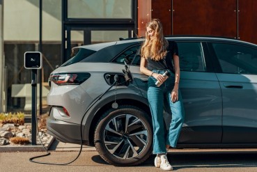 Femme se tenant debout, adossée à un véhicule électrique branché à une borne de recharge.